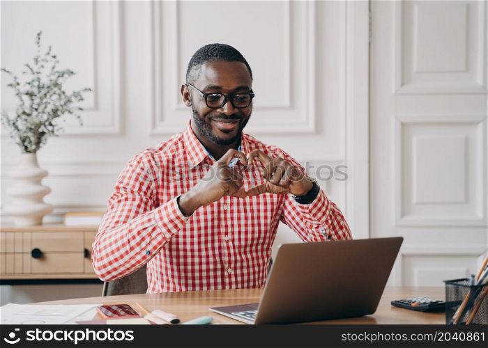 Young happy romantic African man looking at laptop during video call with girlfriend, showing heart symbol with hands and fingers, cheerful employee speaking with loved one during remote work online. Romantic Aframerican businessman looking at laptop having video call showing heart symbol with hands