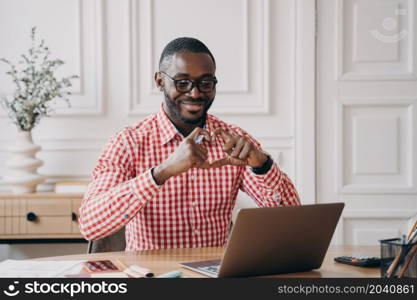 Young happy romantic African man looking at laptop during video call with girlfriend, showing heart symbol with hands and fingers, cheerful employee speaking with loved one during remote work online. Romantic Aframerican businessman looking at laptop having video call showing heart symbol with hands