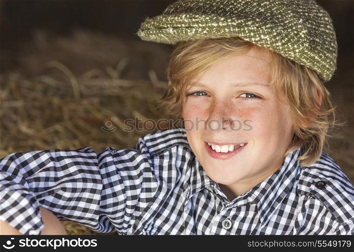 Young happy smiling blond boy child aged about 12 or early teenager wearing a plaid shirt and flat cap sitting on hay or straw bales