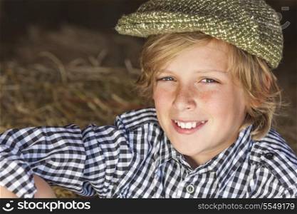 Young happy smiling blond boy child aged about 12 or early teenager wearing a plaid shirt and flat cap sitting on hay or straw bales