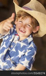 Young happy smiling blond boy child wearing a blue star shirt and cowboy hat sitting on hay or straw bales making a finger gun