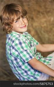 Young happy smiling boy wearing a plaid shirt and sitting on bales of hay or straw