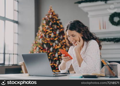 Young happy spanish businesswoman using smartphone while working laptop at home on Christmas, sitting at desk against fir xmas tree with lights, reading sms message on mobile during winter holidays. Young happy spanish businesswoman using smartphone while working laptop at home on Christmas