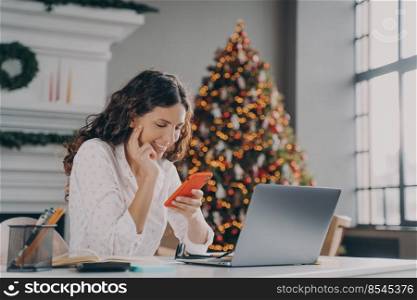 Young happy spanish businesswoman using smartphone while working laptop at home on Christmas, sitting at desk against fir xmas tree with lights, reading sms message on mobile during winter holidays. Young happy spanish businesswoman using smartphone while working laptop at home on Christmas