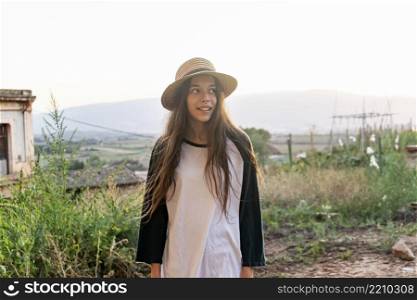Young happy teenager girl with summer clothes and hat standing in a street outdoors while looking away
