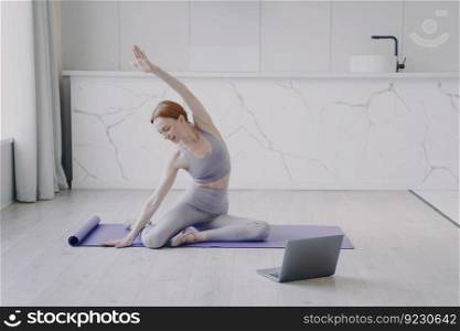 Young happy white woman is stretching and practicing yoga on mat. Concept of e-learning and home online video classes. Laptop is on floor. Sport online and healthy lifestyle on quarantine.. Young woman is stretching and practicing yoga on floor. Sport and healthy lifestyle on quarantine.