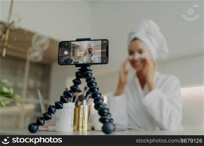 Young happy woman beauty blogger in bathrobe recording video tutorial for skin care blog about using cosmetic under eye patches pads to deal with wrinkles and dark circles, focus on smartphone screen. Smartphone screen with female beauty blogger in bathrobe recording video about skin care for her vlog