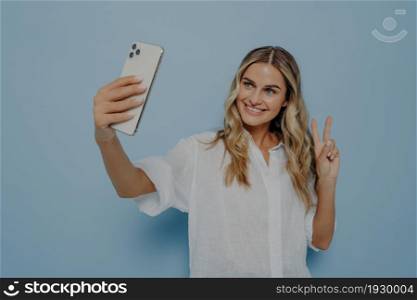 Young happy woman in casual clothes taking selfie while showing peace gesture with her hand, smiling with toothy smile for followers and expressing herself, isolated over blue wall. Blonde teenage girl showing peace sign while taking selfie on smartphone