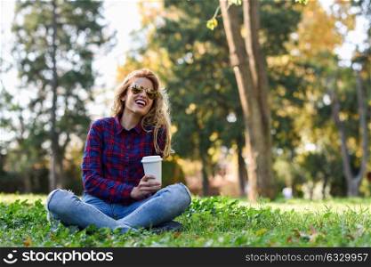 Young happy Woman in checkered shirt and blue jeans with toothy Smile and sunglasses. Blonde girl drinking coffee in park sitting on grass wearing casual clothes smiling