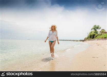 Young happy woman in shirt and hat walking in the sea waves on beach
