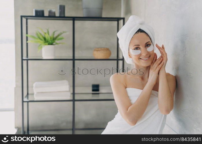 Young happy woman is relaxing at luxurious spa resort. Girl applies anti wrinkle eye patches. Morning beauty routine at home. Attractive european woman wrapped in towel after bathing.. Young happy woman is relaxing at luxurious spa resort. Girl applies anti wrinkle eye patches.
