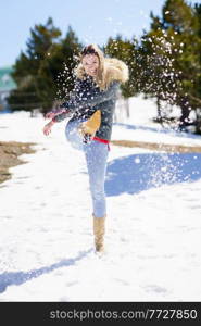 Young happy woman kicking snow in a snow-covered forest in the mountains in Sierra Nevada, Granada, Spain. Female wearing winter clothes playing with snow.. Young happy woman kicking snow in a snow-covered forest in the mountains