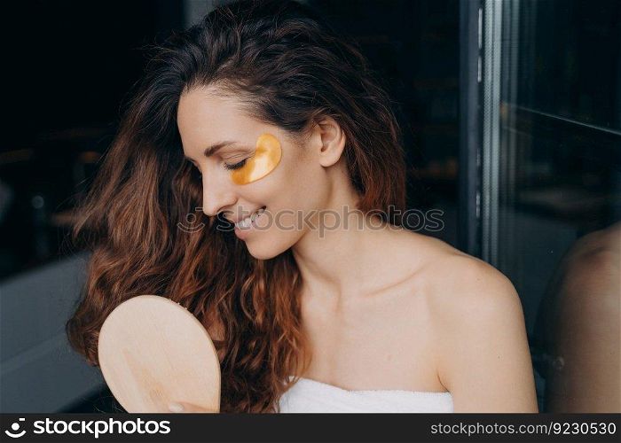 Young happy woman takes shower at home and doing daily hair and face care. Sweet brunette woman combs her soft curly hair with brush. Girl applying eye patches after bathing. Weekend morning at home.. Young happy woman doing daily hair and face care. Woman combs her hair and applies eye patches.