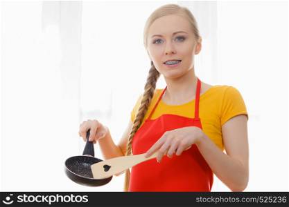 Young happy woman wearing apron holding cooking pan and wooden spatula with heart shape. Female loves cooking. Woman holding cooking utensils