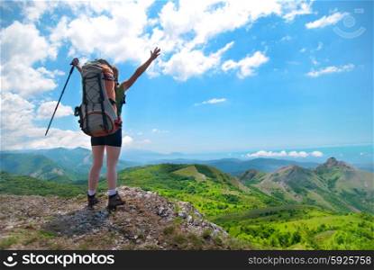 Young happy woman with hiking stick, backpack standing on the rock with raised hands and looking to green landscape