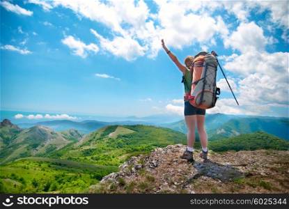 Young happy woman with hiking stick, backpack standing on the rock with raised hands and looking to green lanscape