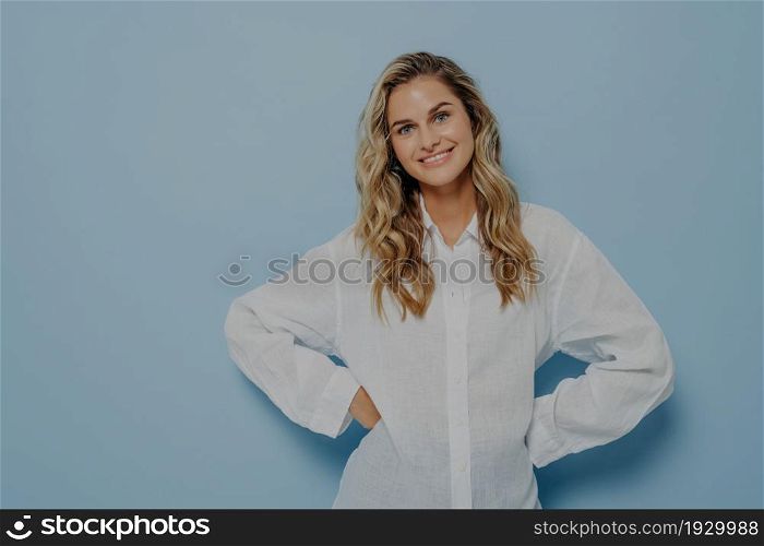 Young happy woman with long wavy hairstyle standing with hands on waist, slightly tilting her head and smiling happily, isolated over blue studio background. Positive human emotions and feelings. Young woman smiling happily while standing with hands on waist and smiling