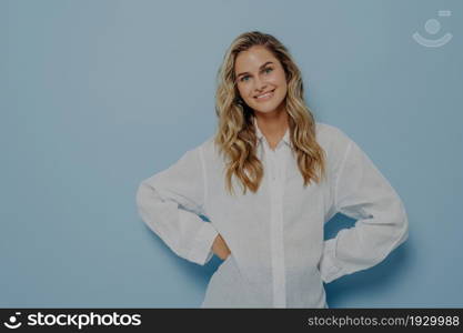 Young happy woman with long wavy hairstyle standing with hands on waist, slightly tilting her head and smiling happily, isolated over blue studio background. Positive human emotions and feelings. Young woman smiling happily while standing with hands on waist and smiling