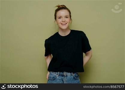Young happy young woman with hair in bun dressed in casual clothes standing in relaxed posture and looking at camera with wide smile on face while posing in studio on green background. Young happy woman in casual clothes expressing positivity