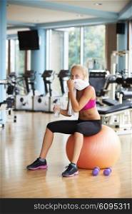 young healthy blonde woman drinking water in fitness gym while sitting on pilates ball