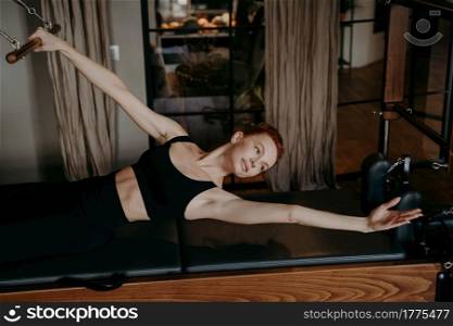 Young healthy female in tight black sportswear lying in stretching position on cadillac reformer table during pilates training in gym. Concept of healthy lifestyle and fitness equipment. Young female pilates instructor lying in stretching arm position on cadillac reformer