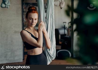 Young healthy fit woman with red hair in bun standing in fitness studio and holding for grey hammock, looking into camera during fly fitness practice, being ready for antigravity yoga exercises. Young woman being ready for antigravity yoga exercises in studio, looking in camera