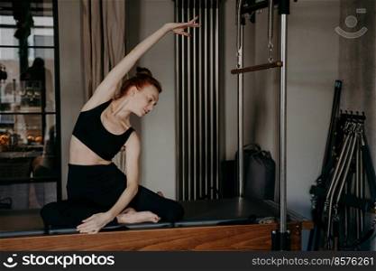 Young healthy woman doing pilates exercises on cadillac reformer, beautiful slim sports trainer showing flexibility and stretching muscles in black sportswear. People and fitness, wellness concept. Young ginger woman doing pilates exercises with reformer
