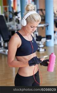 young healthy woman drinking water in fitness gym while sitting on pilates ball and listening music on headphones from smartphone