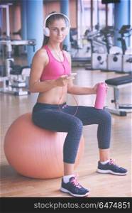 young healthy woman drinking water in fitness gym while sitting on pilates ball and listening music on headphones from smartphone. woman with headphones in fitness gym