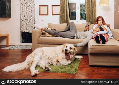 Young heterosexual couple together on sofa with their dog using smartphone and drinking from a cup