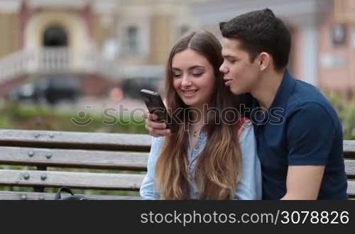 Young hipster couple watching video content on mobile phone while sitting on the bench embracing each other in park. Cheerful couple surfing the net with smartphone together and talking while relaxing outdoors during romantic date. Closeup.