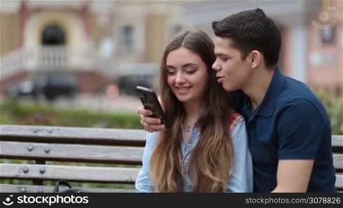 Young hipster couple watching video content on mobile phone while sitting on the bench embracing each other in park. Cheerful couple surfing the net with smartphone together and talking while relaxing outdoors during romantic date. Closeup.