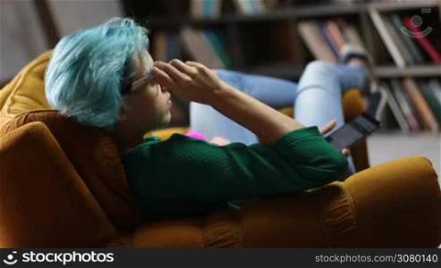 Young hipster girl with blue hair and eyeglasses sitting in comfortable chair using modern smartphone device at home. Trendy young woman with mobile phone communicating with friends in social networks. Freelancer working from home via cellphone.