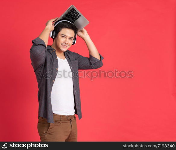 Young hipster handsome asian guy with wireless earphones on head playing games on laptop computer while standing isolated on red Teenager listening to music via headphones during work on notebook