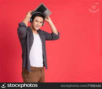 Young hipster handsome asian guy with wireless earphones on head playing games on laptop computer while standing isolated on red Teenager listening to music via headphones during work on notebook
