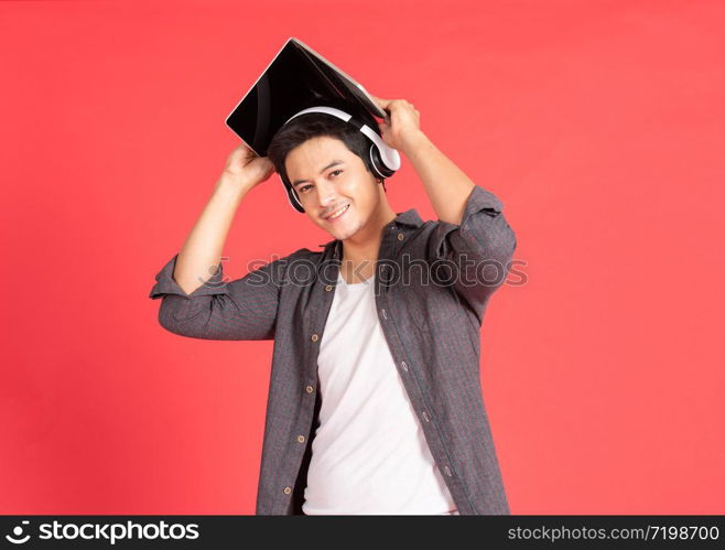 Young hipster handsome asian guy with wireless earphones on head playing games on laptop computer while standing isolated on red Teenager listening to music via headphones during work on notebook