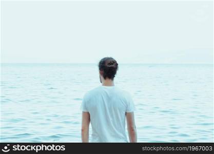 Young hipster male with long hair and a bun looking to the ocean, during a sunny day, reflection and yoga concept, serenade, copy space