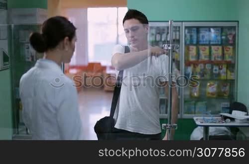 Young hipster man with pain in back discussing over medicines with pharmacist in pharmacy. Positive woman pharmacist wearing uniform assisting customers in pharmaceutical shop. Female druggist suggesting medical drug to patient at local drugstore.