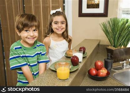 Young Hispanic brother and sister sitting at kitchen bar with healthy breakfast smiling at viewer.