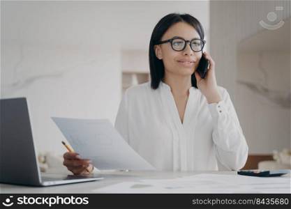 Young hispanic businesswoman in glasses has phone talk. Girl is working remote at home office on quarantine. Manager, accountant or business assistant is doing paperwork using laptop, examining data.. Young businesswoman in glasses has phone talk. Girl working remote at home office, doing paperwork.