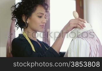 Young hispanic female dressmaker adjusting clothes on tailoring mannequin and smiling at camera. Dolly shot