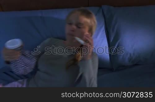 Young hispanic girl in pajamas, laying on bed with fever and ice bag on head, measuring temperature with thermometer at night. High angle view, slow motion 60p