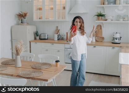 Young hispanic woman boasting with her stylish kitchen. Housewife has video phone call at home. Girl shows modern white scandinavian interior into camera. Stove, worktop and cuisine.. Young hispanic woman boasting with her stylish kitchen. Housewife has video phone call at home.