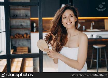 Young hispanic woman combs her long curly hair with brush. Relaxed smiling girl wrapped in towel after bathing. Modern interior of apartment. Relaxation, grooming and p&ering at home concept.. Young hispanic woman combs her long curly hair. Relaxation, grooming and p&ering at home.