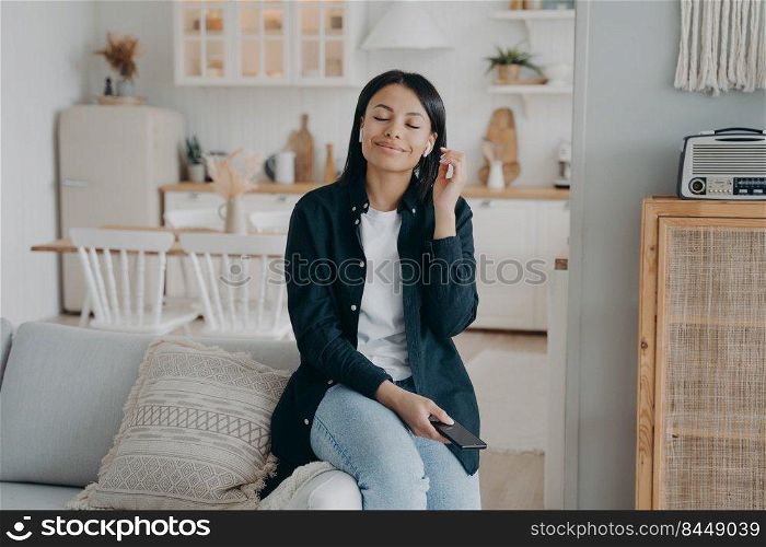 Young hispanic woman enjoying music listening in airpods. Girl relaxing with her eyes closed. Favourite romantic song listening and dreaming. Lady is sitting on couch and having nice weekend morning.. Young hispanic woman enjoying music listening in airpods, relaxing with her eyes closed.