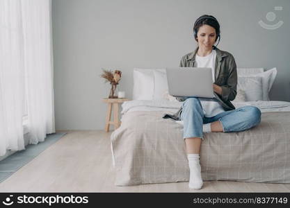 Young hispanic woman has group call in earphones. Freelancer has internet session. Online lesson or meeting on quarantine. Girl staying home and sitting on bed in bedroom. Modern white interior.. Young hispanic woman has group call in earphones. Freelancer has internet session at home.