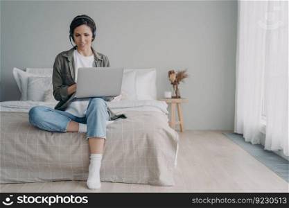 Young hispanic woman has group call in earphones. Freelancer has internet session. Online lesson or meeting on quarantine. Girl staying home and sitting on bed in bedroom. Modern white interior.. Young hispanic woman has group call in earphones. Freelancer has internet session at home.