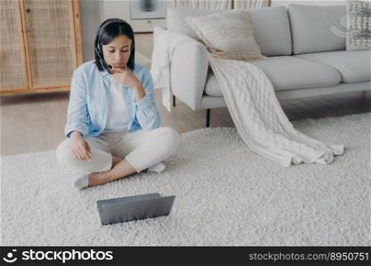 Young hispanic woman in earphones with microphone has internet session on laptop. European girl is listening to video. Distant learning from home. Virtual meeting and communication on quarantine.. Young woman in earphones with microphone has internet session and listening to video at home.