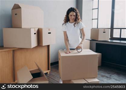 Young hispanic woman is packing containers with sticky tape. Happy mover is wrapping cardboard boxes with packing tape. Moving service worker preparing boxes for shipping and storage.. Young hispanic woman is packing containers with sticky tape. Happy mover wrapping cardboard boxes.