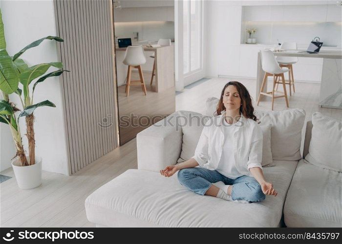 Young hispanic woman is practicing yoga and meditation at home. Girl in white is sitting on couch with her eyes closed. Posture exercise, lotus asana, gymnastics. Wellbeing and morning routine.. Young hispanic woman is practicing yoga and meditation at home. Wellbeing and morning routine.